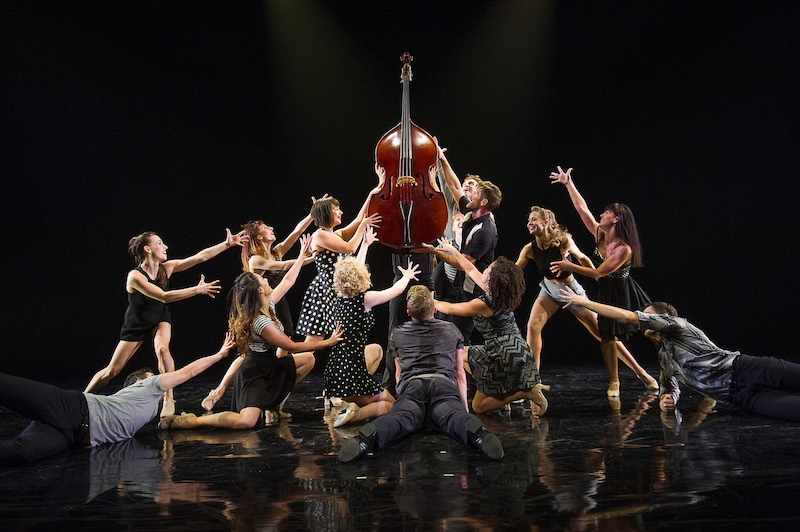 A group of dancers in white and back huddle around a stand up bass. Their arms outstretched around it.